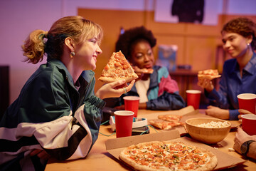 Side view portrait of smiling girl eating pizza with group of friends at house party 80s style,...