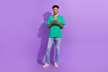 Fototapeta premium Full size cadre of young smiling blogger guy wear green t-shirt denim jeans hold phone browsing twitter isolated on purple color background