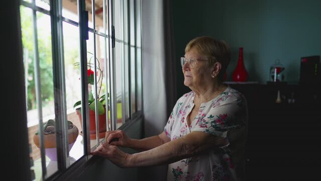 Elderly Woman with thoughtful Expression Standing by Window at Home looking out in solitude, Lonely Contemplative Scene of an Older Person in authentic domestic lifestyle