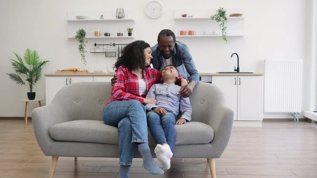Portrait of multiracial family posing together on soft couch in open-plan kitchen on sunny day. Cheerful mother and cute son snuggling into father's arms enjoying comfort and security.