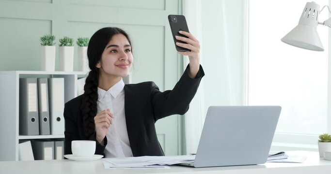 Indian businesswoman, radiates confidence and positivity. With a warm beverage in hand, she grabbing her smartphone to capture a cheerful selfie. She have a break from work.