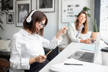 Joyful lady office manager in headphones enjoying music and singing while working in office with...