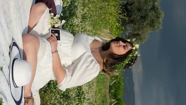 In slow motion, a pregnant woman in a white dress and daisy crown shares a tender moment, displaying her baby's ultrasound image, while seated on a picnic blanket amidst greenery.