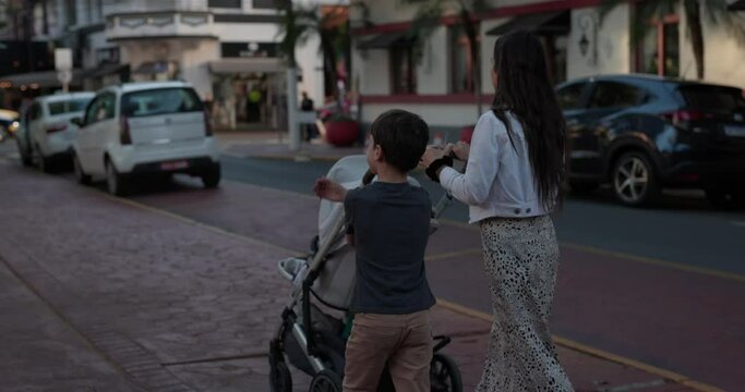 Young mom walks down tropical south american street at sunset with son while pushing stroller