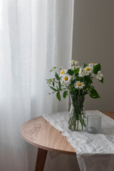 A bouquet of daisies and jasmine in a glass vase on a round wooden table. Summer interior concept