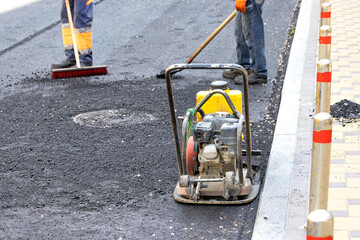 An old compacting vibrating plate stands at the curb against the backdrop of a working brigade of road workers.