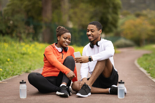 Sports blog, app for body care. Glad young black couple sit on road, talking, relaxing with bottles of water