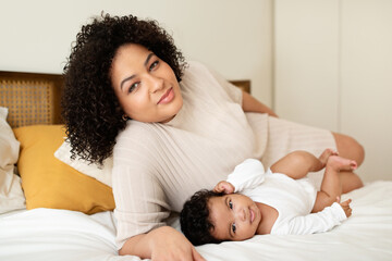 Obraz na płótnie Canvas Mom and newborn kid. Positive black millennial lady lies with little baby on white bed, enjoys comfort