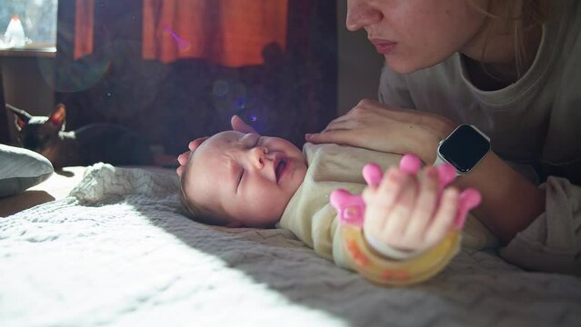 Caucasian mother trying to calm his crying newborn baby girl laying on the bed. Mom stroking upset little child. Indoor, cat in background. Mommy's care, growing, parenting