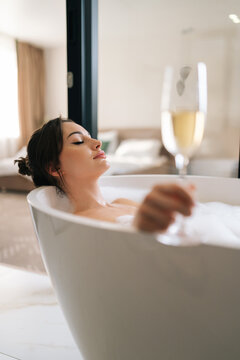 Vertical side view of young happy woman lying closed eyes in luxury white bathroom holding glass of champagne. Closeup of gorgeous female relaxing in bath during luxurious recreation for weekend..