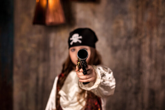 Little girl actress 7-8 year old in pirate's image shooting pistol in camera at gray textured wall, serious gun. Kid in pirate corsair costume on stage. Theatre performance concept. Copy ad text space