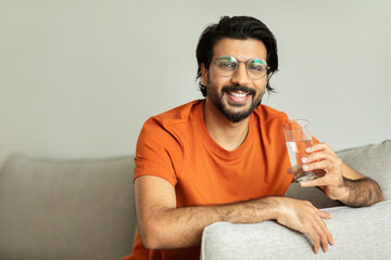 Cheerful millennial middle eastern guy in glasses with glass of water, sits on sofa, enjoy lifestyle in living room