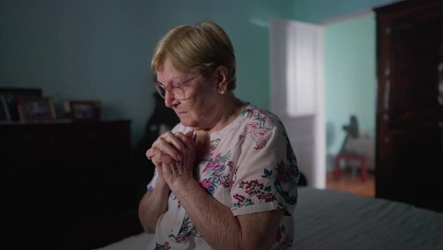 Religious Elderly woman sitting by bedside in Prayer. HOPEFUL older lady Praying to GOD in bedroom, authentic real life domestic lifestyle depicting old age in FAITH