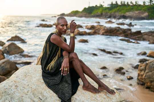 Androgynous ethnic fashion model in luxury dress, jewelry sits on rocks by ocean. Gay black person in jewellery, posh clothes poses gracefully in tropical seaside location. Pride LGBTQ bipoc concept.