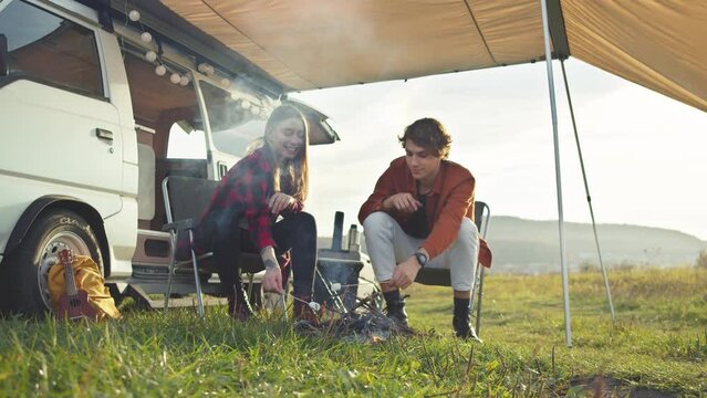 Young caucasian couple roasting marshmallows on campfire, talking and smiling. Hipster girl and boy take a nature retreat. Romantic road trip, outdoor weekend, happy picnic in fresh air