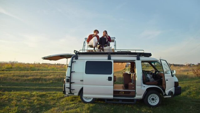 Nice view of young couple sitting on rooftop white camper van. Countryside background, blue sky, green grass. Romantic road trip to backcountry, nature landscape, outdoor weekend