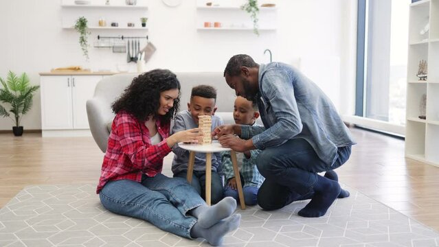 Four-person family playing board game with wooden blocks on small table in living room. Happy people being engaged in enjoyable activity showing importance of love and care.
