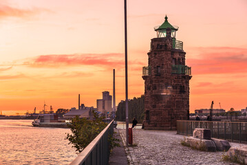 Stunning summer sunset over a historic old stone lighthouse at the end of a cobblestone pier on a river harbour - obrazy, fototapety, plakaty