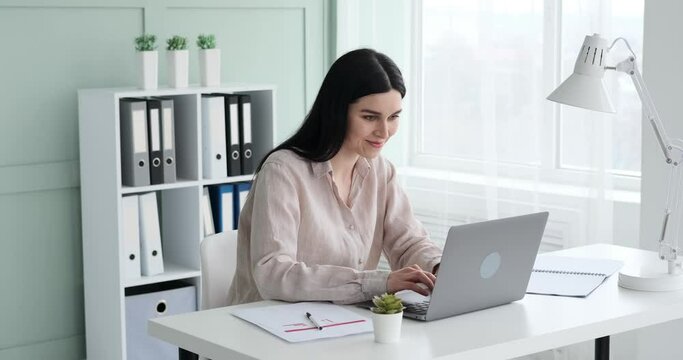 Cheerful businesswoman wearing a shirt sitting at her office desk, typing on a laptop, and smiling widely with her teeth showing. She seems to be in a great mood and enjoying her work.