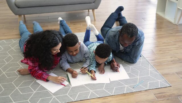 Top view of talented multiracial couple coloring pictures together with focused children while staying at home. Thoughtful parents developing artistic skills of little sons after walk outdoors.