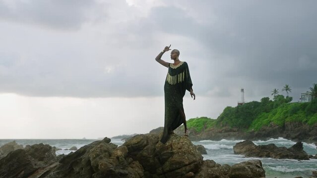 Androgynous person of color stands in boutique posh dress on rock above ocean coastline. Trans sexual ethnic fashion model in epic outfit wears golden jewellery, posing like Oshun goddess concept.