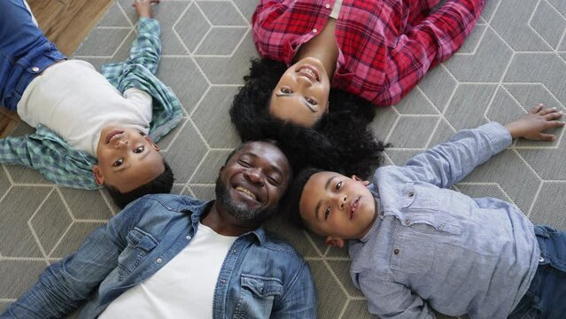 Top view of multiethnic four-person family lying in circle with heads together on floor. Smiling parents and joyful sons sharing enormous positive energy in home interior.