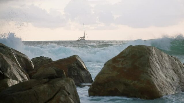 Yacht ship sailing at stunning sea view at sunset, waves break against big rocks and splash. Breathtaking ocean sunset on rocky beach, rough water. Sailboat on background of meditative seascape.