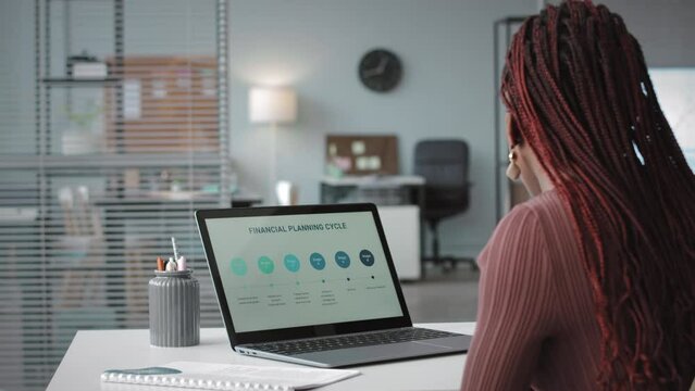 Back view of young African American woman analyzing financial planning cycle on laptop and doing paperwork while working in office