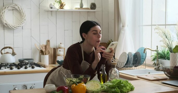 Caucasian woman in apron at kitchen, standing near countertop, with a phone in her hand, engrossed in its screen, while savoring a refreshing salad from a bowl.