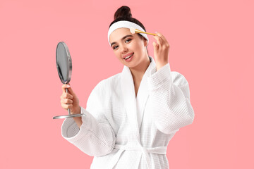 Naklejka premium Pretty young woman in bathrobe applying makeup on pink background