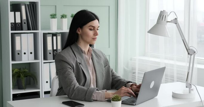 A confident and focused businesswoman wearing a shirt and jacket sits at desk, typing on laptop with a light smile on her face. She looks comfortable and in control, getting her work done efficiently.
