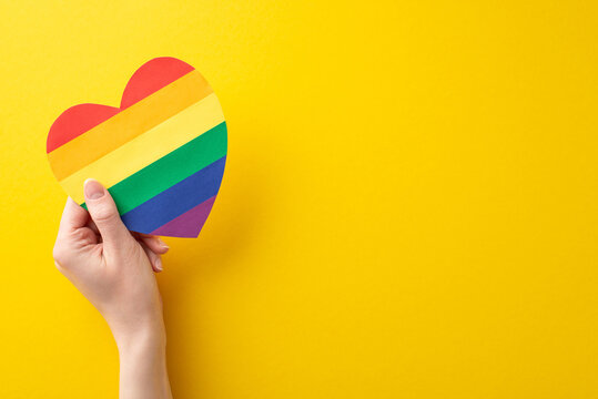 LGBT History Month theme: first person top view of young woman's hand holding rainbow heart postcard on vivid yellow background with blank space for text or advert