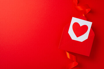 Top view photo of empty space and envelope with the red heart inside surrounded by bright red ribbon and paper origami hearts on isolated red background with copy-space