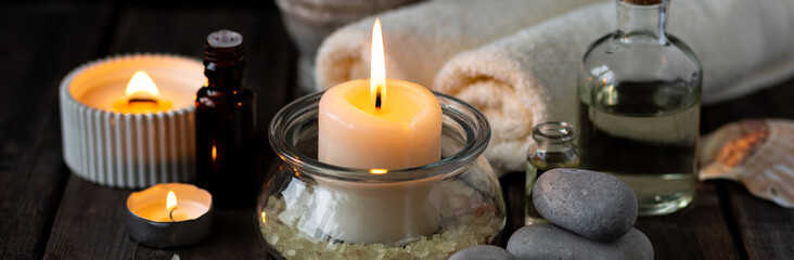 Obraz na płótnie Canvas Spa setting with essential oil, candle, sea salt, pebbles, towel on dark wooden background. Massage, aromatherapy. Natural organic ingredients for relaxation, detention, wellness. Banner copy space