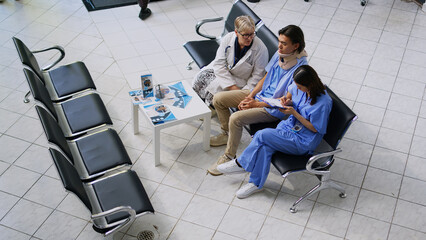 Fototapeta Elderly doctor examining patient with neck injury while asian nurse writing medical expertise on clipboard during checkup visit in hospital lobby. Young man wearing cervical collar in waiting area obraz