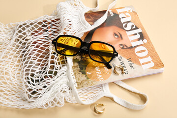Stylish sunglasses and string bag with magazine on beige background