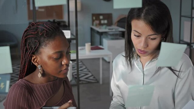 Chest up of two young creative multiethnic female colleagues putting sticker notes on glass window in office while brainstorming ideas and discussing business plan