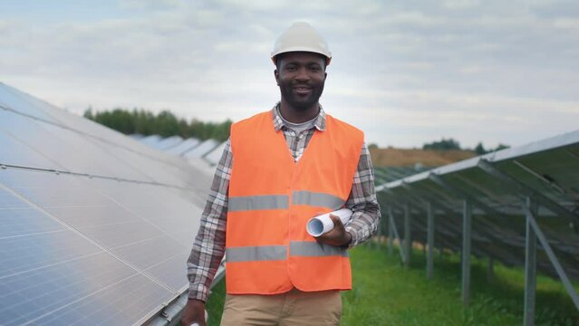 Portrait of industrial worker on solar panel background outside. African American employee dressed in protective vest and hardhat standing with drawing on paper. Concept of green electricity.