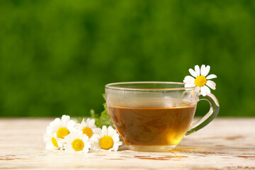 Glass cup of tea with beautiful chamomile flowers on white wooden table outdoors