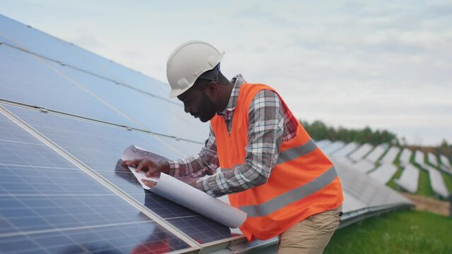 Portrait of an African-American employee working in the production of solar panels. Worker wearing protective vest and hard hat looks and examine blueprint outside. Concept of green electricity.