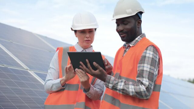 Male and female workers on background of solar panels. African American engineer wearing in hard hat and reflective jacket at workplace. Woman holds tablet and explain tasks to colleague.