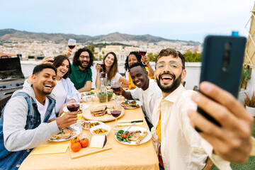 Young group of friends gathered sitting around a table enjoying a barbecue lunch. Multiracial people taking a selfie while eating meat and drinking wine at summer party celebration. Friendship concept