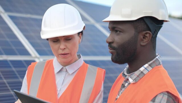Portrait of male and female employees in hard hats and protective vests are working on solar panel background outside. African American engineer using tablet for making tasks.