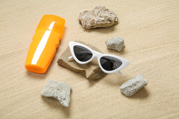 Bottle of sunscreen cream, sunglasses and stones on sand, closeup
