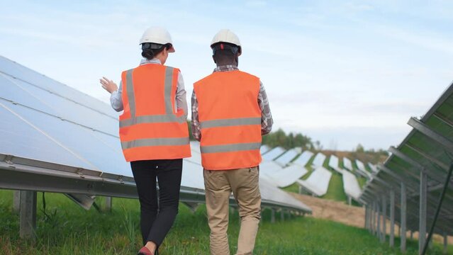 Portrait of male and female employees from the back . African American man and European woman in hard hats and protective vests are walking on solar panel background outside.