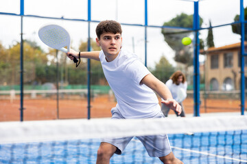 Portrait of sporty young guy playing padel on open court on summer day, ready to hit ball. Sport...