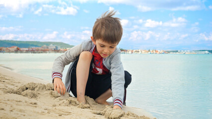Boy sitting on the sea beach, digging and playing with sand. Concept of tourism, travel, summer vacation and weekends