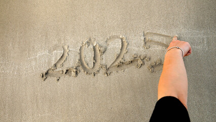 Closeup of young woman writing new year 2025 on the wet sand at sea beach