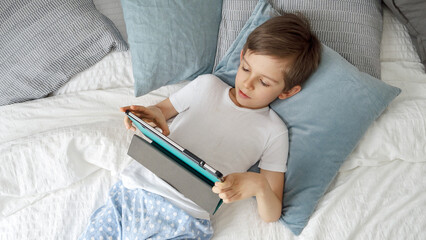 Little boy is lying on his bed, using a tablet computer during the day. Child using gadget, education and development.