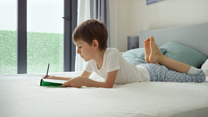 Little boy in pajamas lying on bed at big window and doing homework in notebook. Concept of...